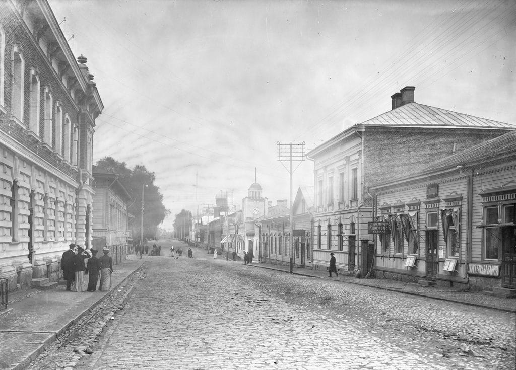 Kaupunkimaisema 1900-luvun alusta Mikkelissä. Mukulakivikatu, jonka molemmin puolin rakennuksia. Muutamia ihmisiä kadulla.