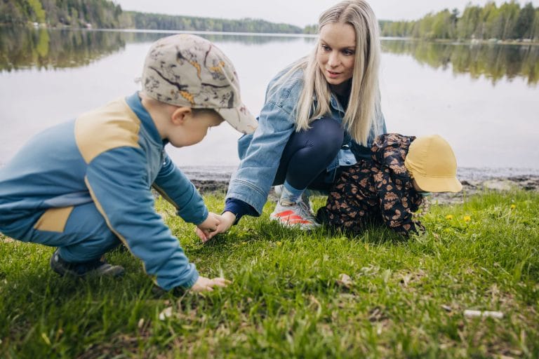 Lapset ja äiti tutkivat nurmikkoa järven rannalla.