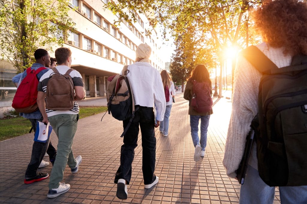 Nuoret kävelevät selkä kameraan päin kadulla kohti puiden takaa loistavaa aurinkoa.