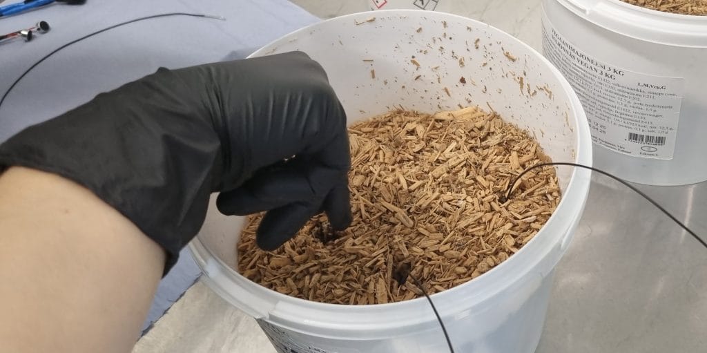 A hand places wires into a bucket partially filled with a material resembling wood chips.