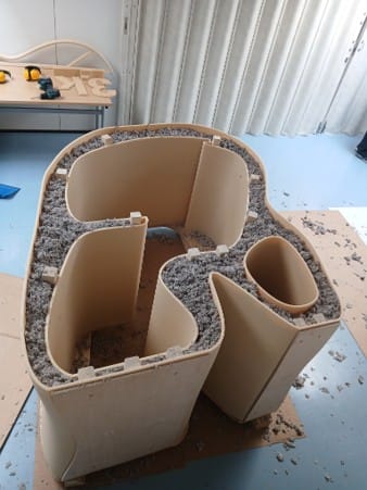Työtila kaadettuna kyljelleen etuseinä lattiaa vasten. 3D-tulostettujen sisä- ja ulkokuorien välissä näkyy eristevillaa ja poikkipuiden päät. 
