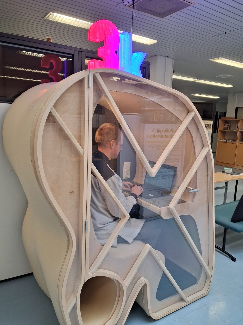 Kaarevamuotoinen 3D-tulostettu hiljainen tila, jossa ihminen istuu ja kannattavan tietokoneen äärellä.