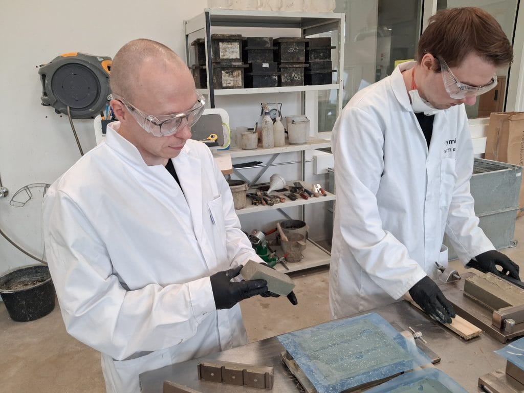 Kaksi henkilöä laboratoriossa tarkastelemassa prismakoekappaleita ja irrottamassa niitä muoteista. Taustalla näkyy betonilaboratorion välineistöä.