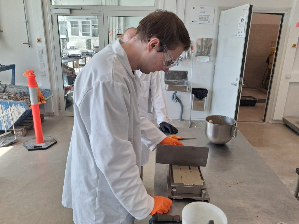 Kaksi henkilöä laboratoriossa tarkastelemassa prismakoekappaleita ja irrottamassa niitä muoteista. Taustalla näkyy betonilaboratorion välineistöä.