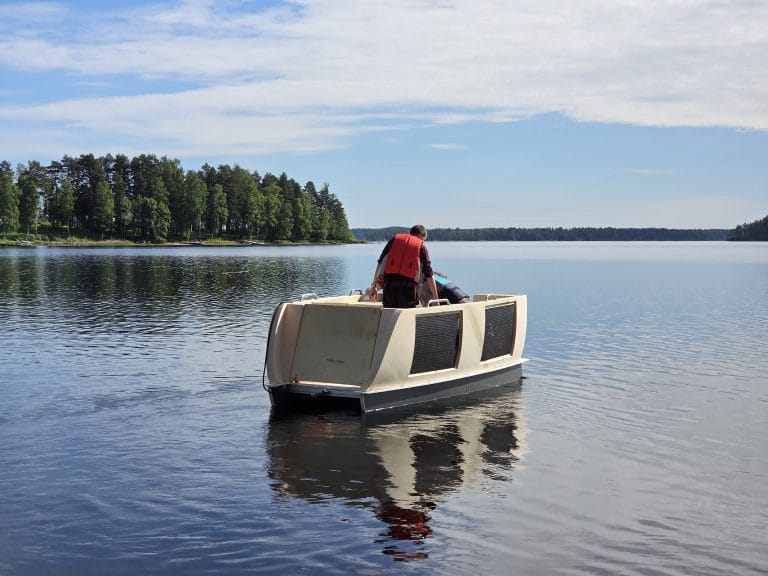 Vedessä alus, jonka keulassa avautuva lastausramppi ja kyljissä aurinkopaneeli. Kyydissä kaksi henkilöä ja taustalla metsää. Aurinkoinen keli.