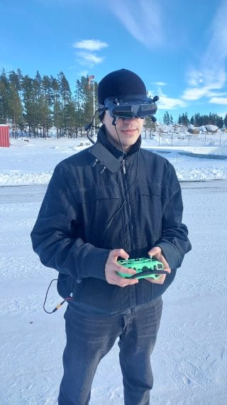 Mies ulkoilmassa talvivaatteet päällä, päässään FPV-lasit ja käsissä droonin kauko-ohjain. Taustalla luminen maisema.