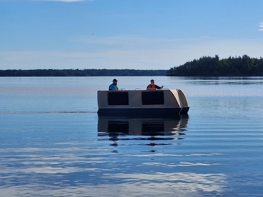Vedessä alus, jonka keulassa avautuva lastausramppi ja kyljissä aurinkopaneeli. Kyydissä kaksi henkilöä ja taustalla metsää. Aurinkoinen keli. 