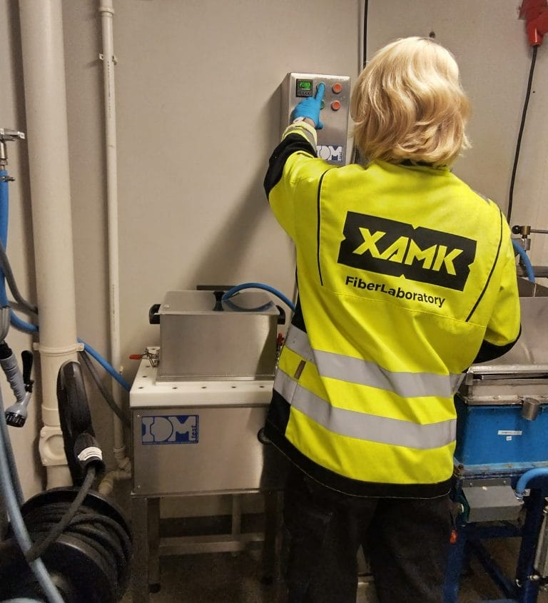 Ihminen keltaisessa liivissä painelemassa laitteen nappeja kuitulaboratoriossa.